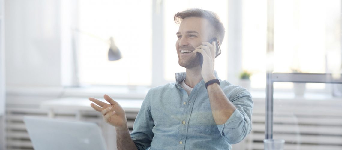 Portrait of handsome young man laughing cheerfully while speaking by phone sitting at desk in office, copy space